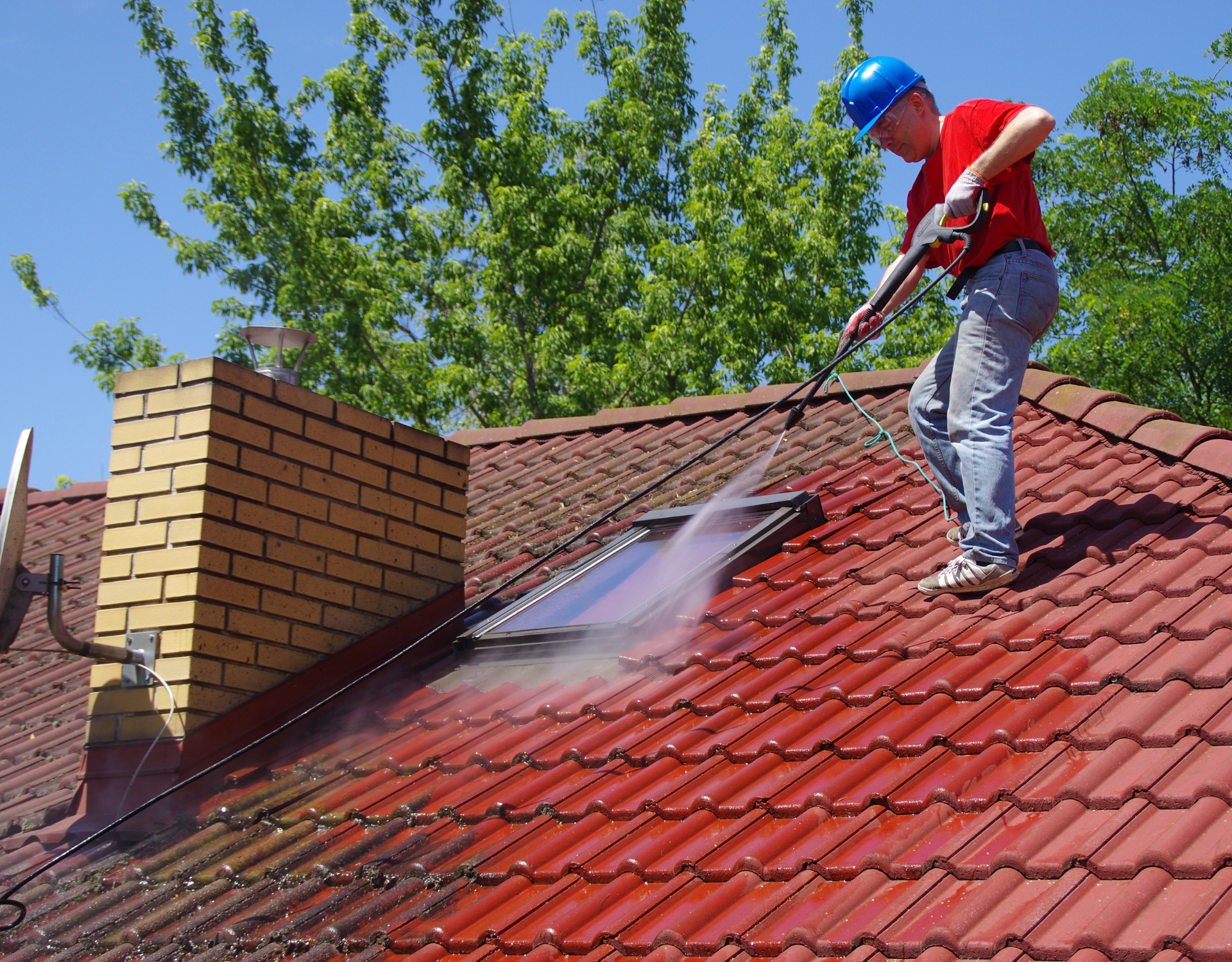 Worker pressure-washing a red tiled roof beside a brick chimney under a blue sky