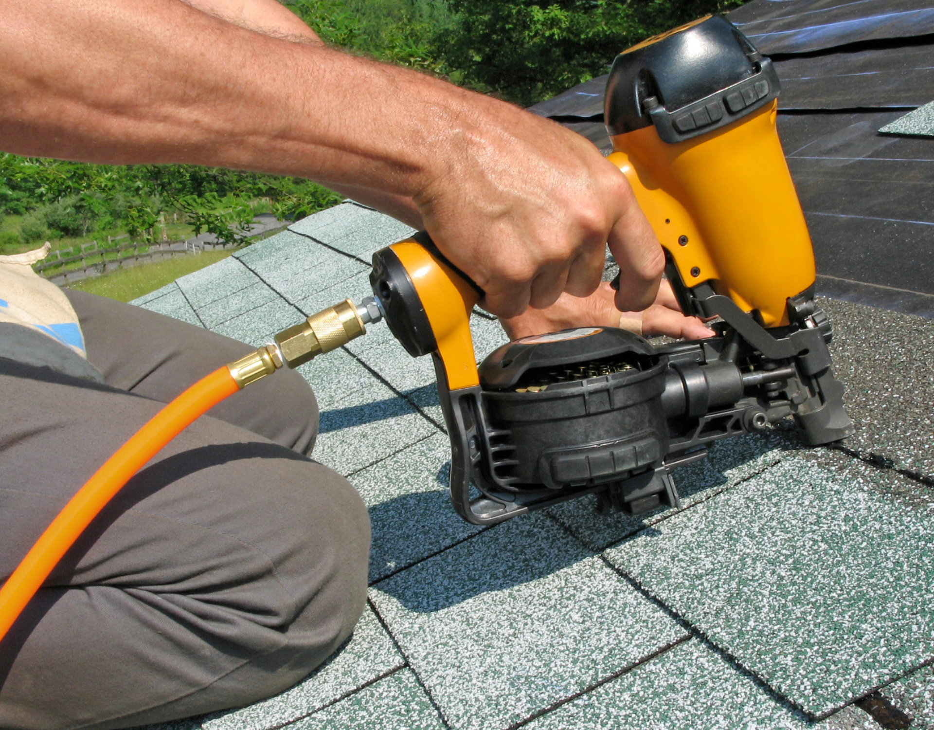 Worker using a yellow roofing nail gun on asphalt shingles