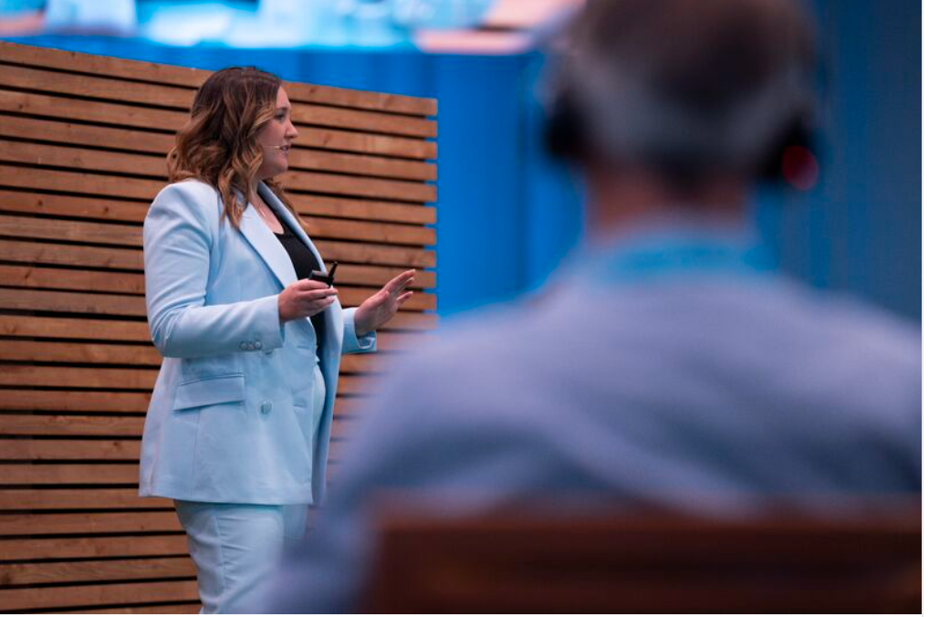 Woman in light blue suit giving a presentation; wood panel backdrop, audience member in foreground.
