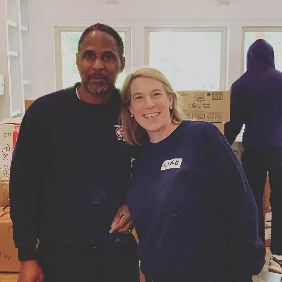 Two people smiling in front of moving boxes; one is African American, one is white. A third person is in the background.