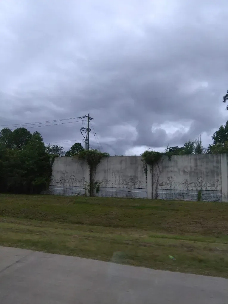 Gray concrete wall under a cloudy sky, with grass and trees in front.