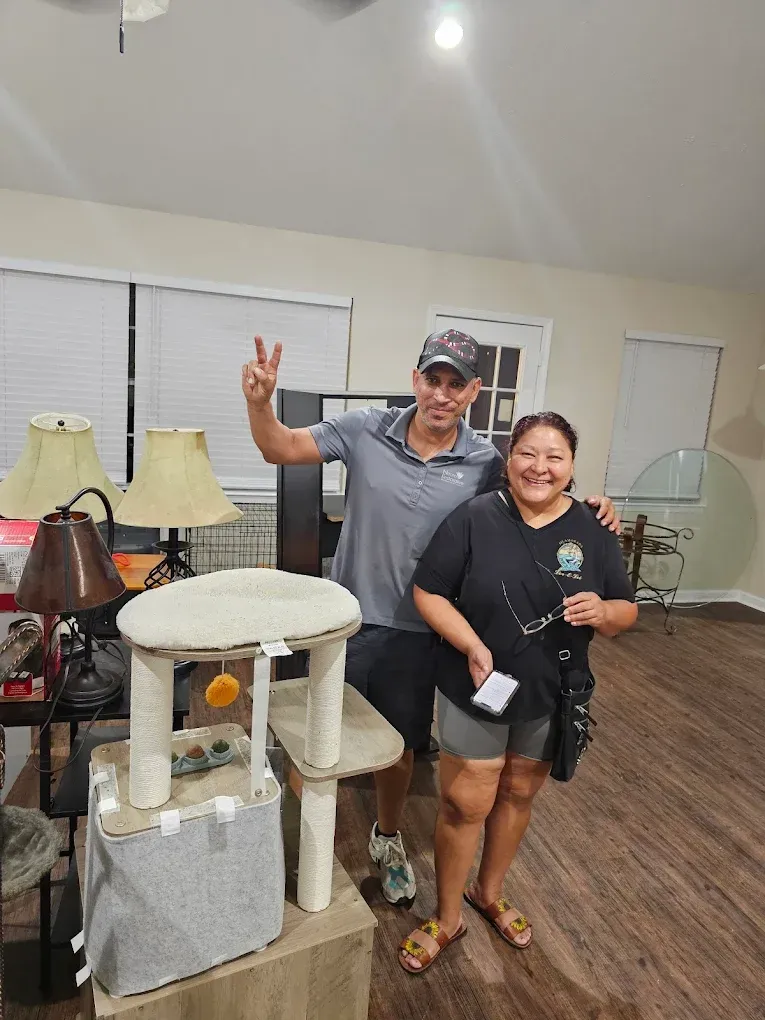 Man gives peace sign next to a woman; they stand near a cat tree in a room with lamps and furniture.