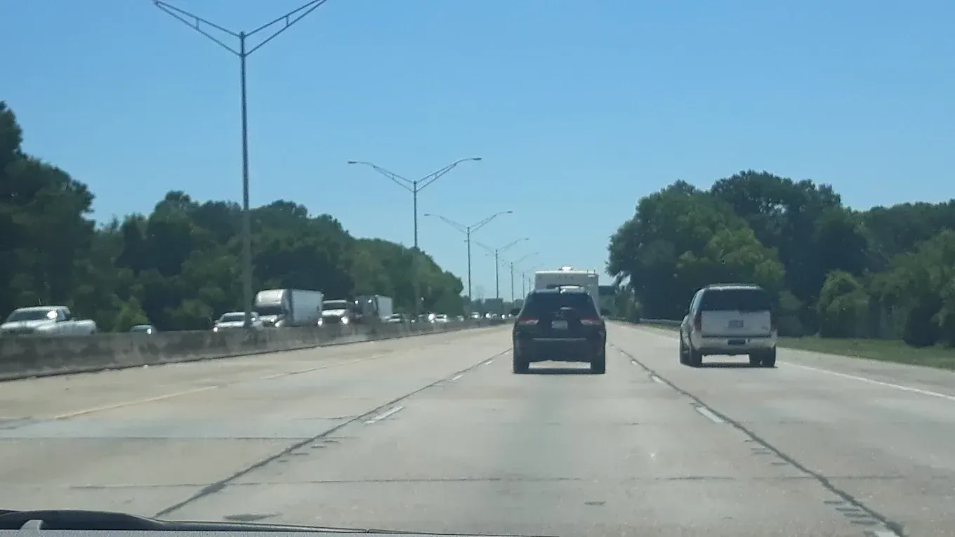 Cars driving on a multi-lane highway, trees line the sides, clear blue sky overhead.