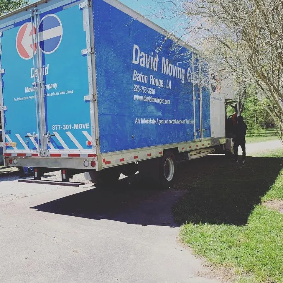 Blue David Moving truck parked on a driveway in Baton Rouge. A person stands near the door.