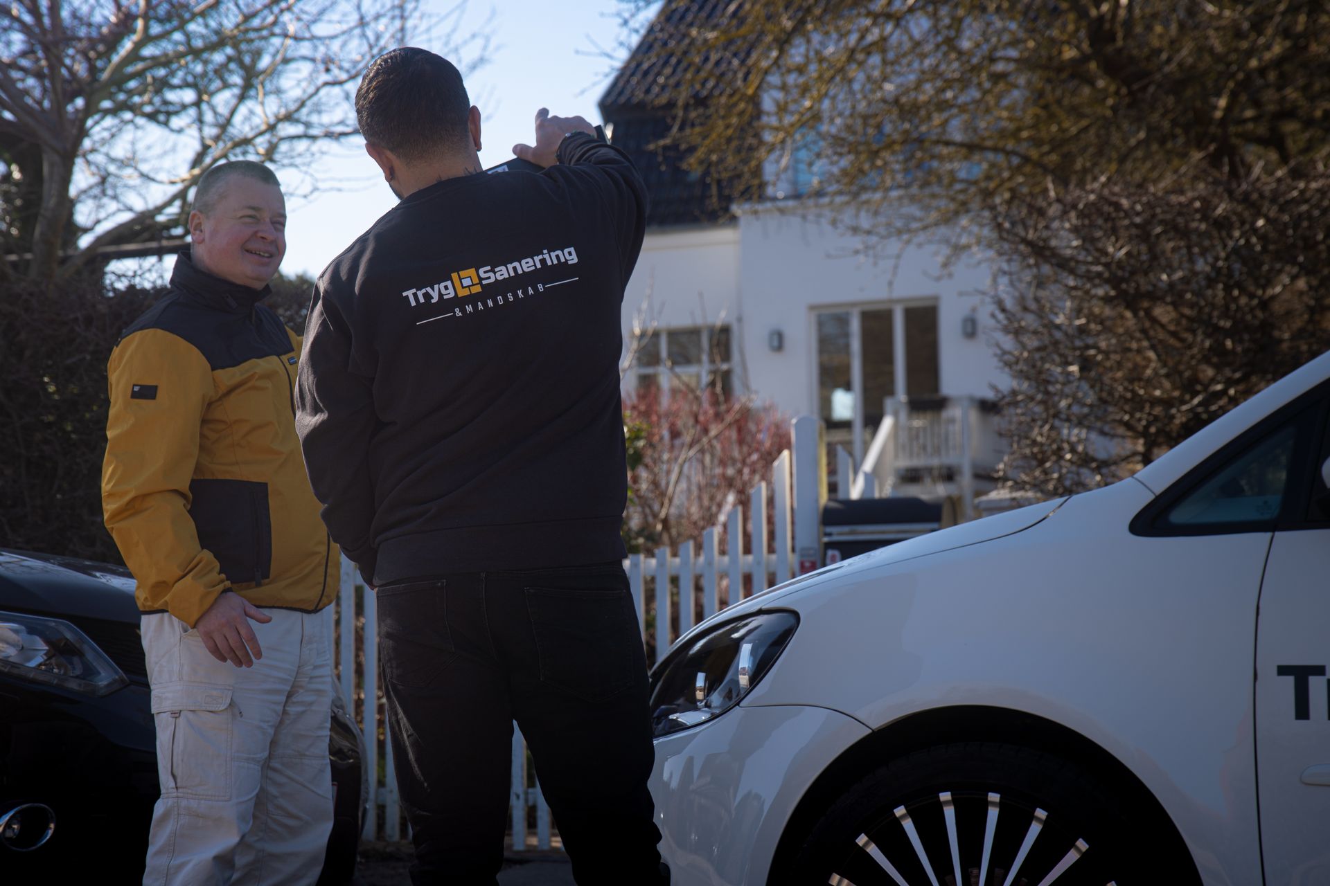 To mænd står ved siden af ​​en hvid bil foran et hus.