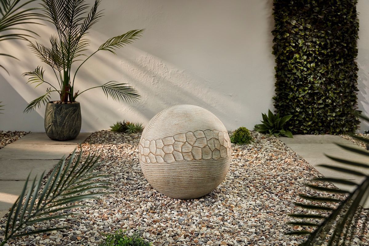 Stone sphere fountain in a gravel bed with greenery and a white wall backdrop.