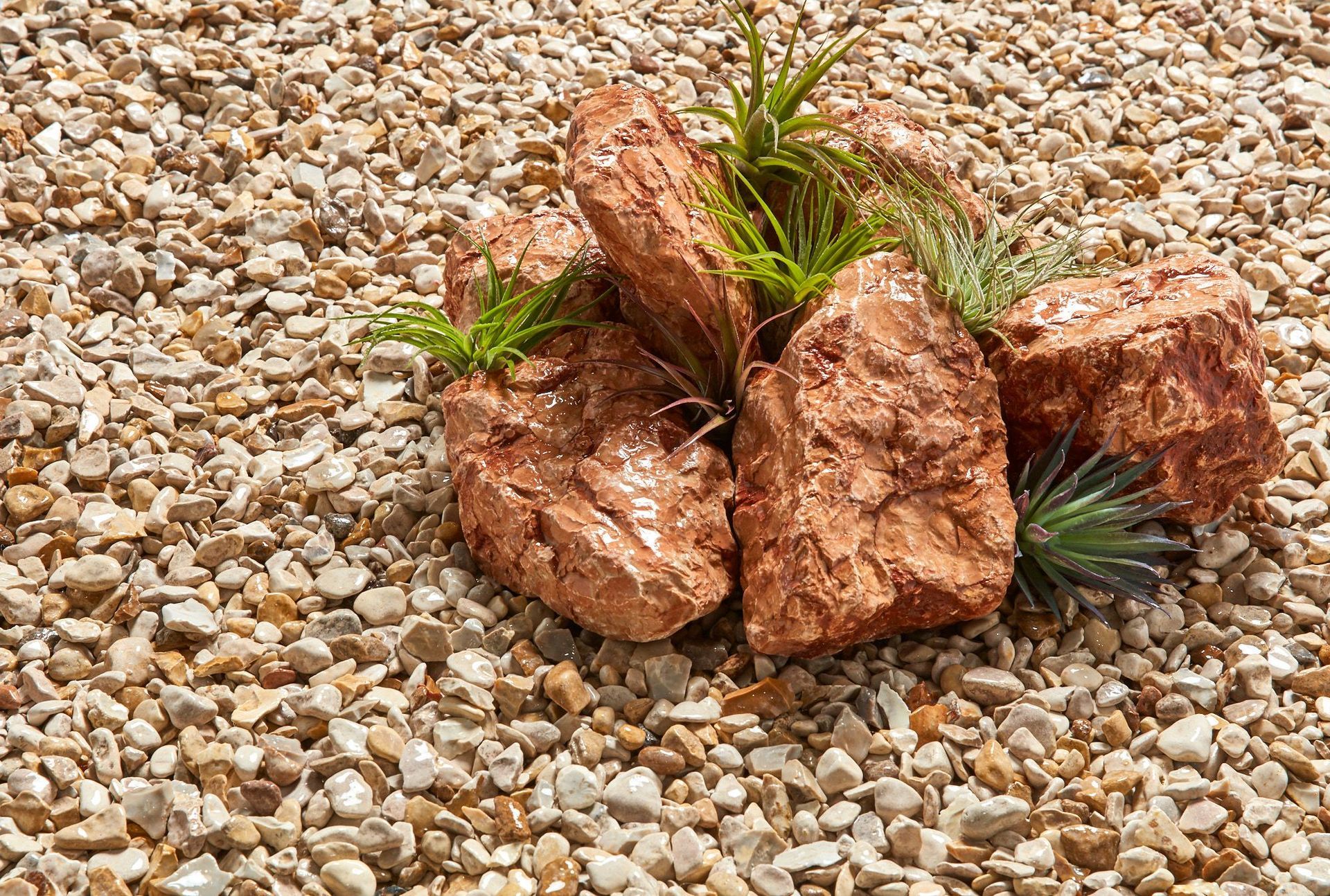 Red rocks with green plants atop gravel.