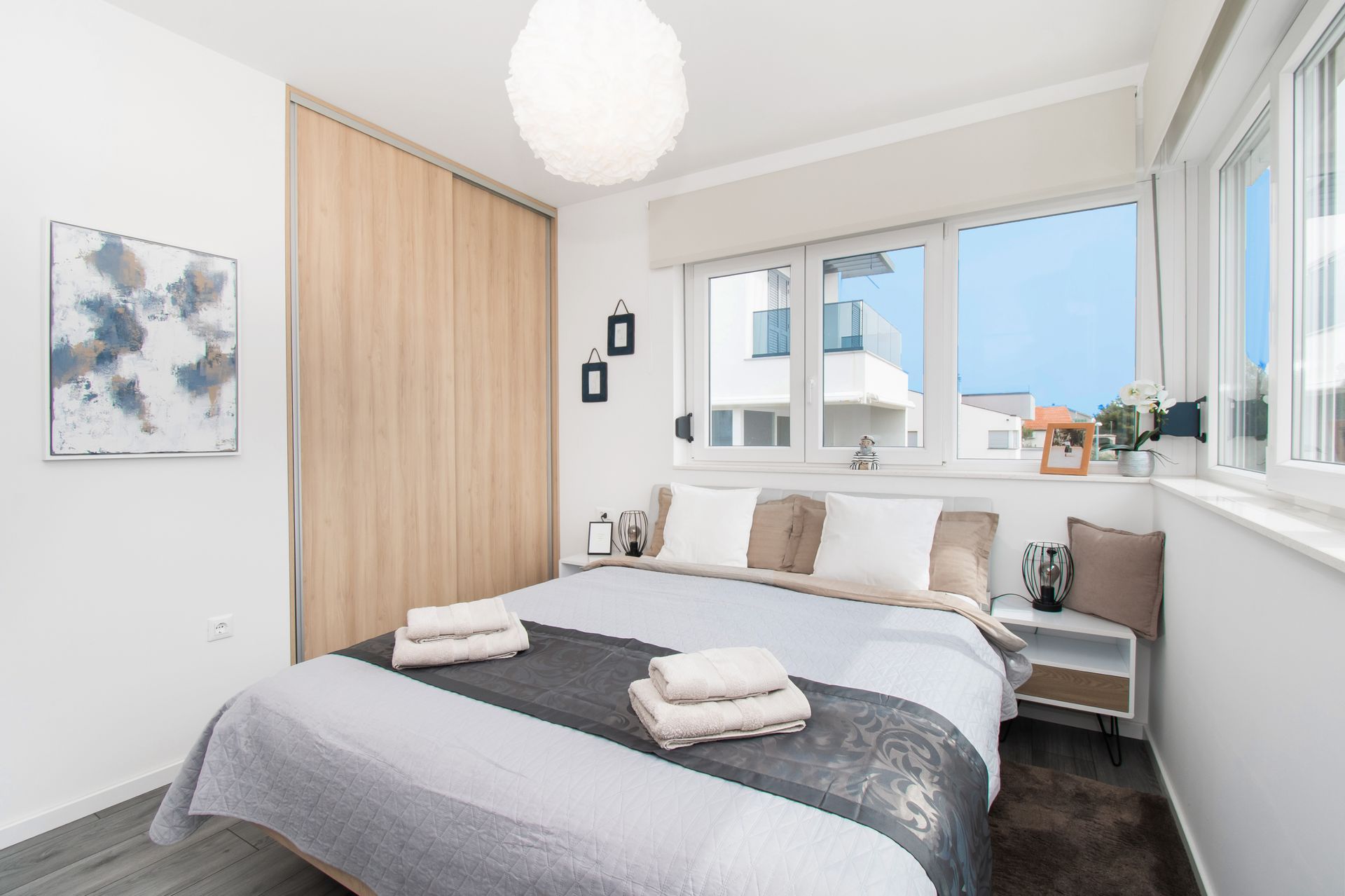 Bedroom with a bed, wooden closet, artwork, and large windows. Sunlight streams through.