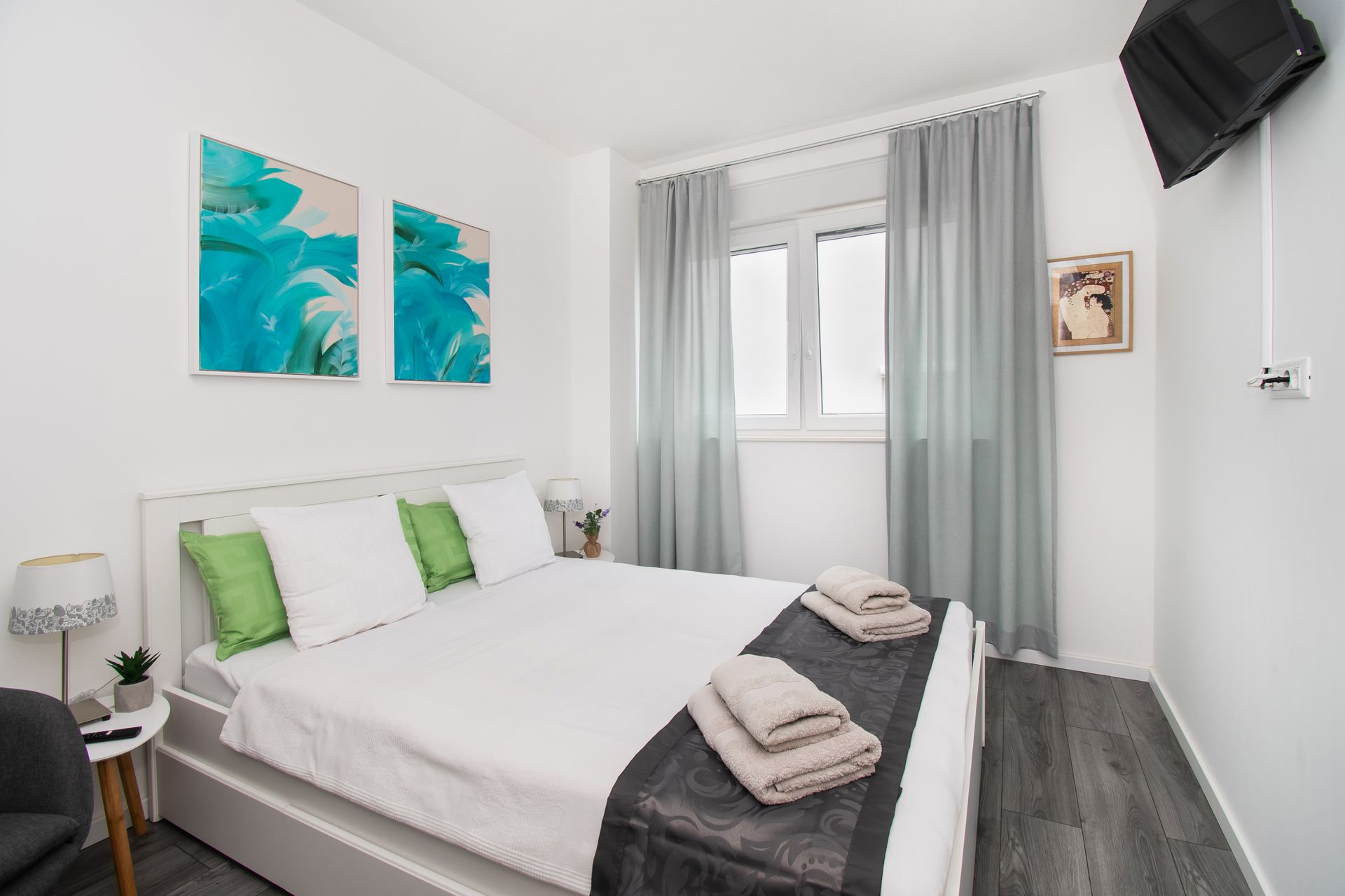 Bedroom with white walls, bed, teal artwork, gray curtains, and a mounted TV.