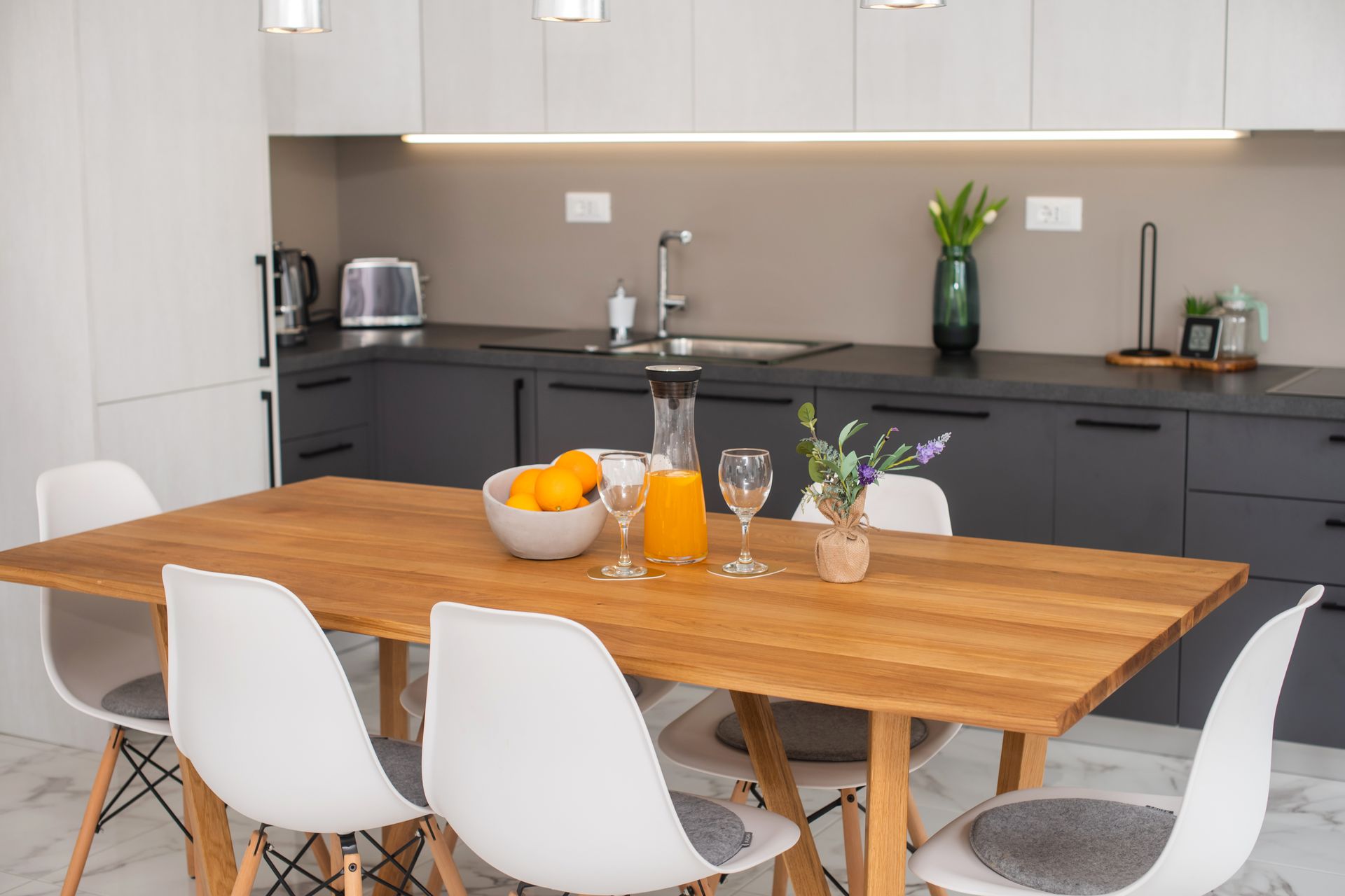 A modern kitchen with a wooden table, six white chairs, and gray cabinets. Oranges and drinks are on the table.