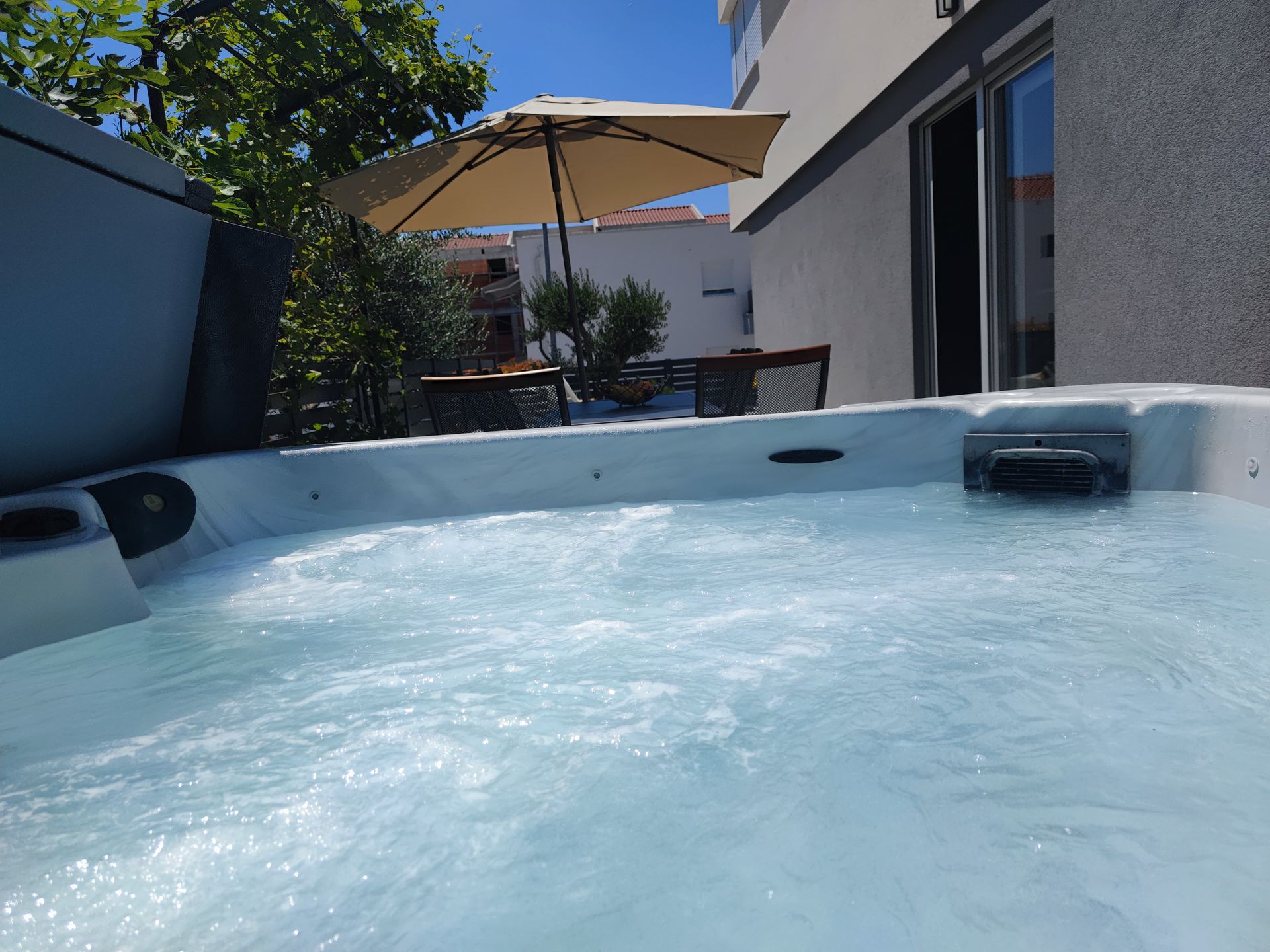 Hot tub bubbling with water, patio with umbrella, table, and chairs. Sunny day.