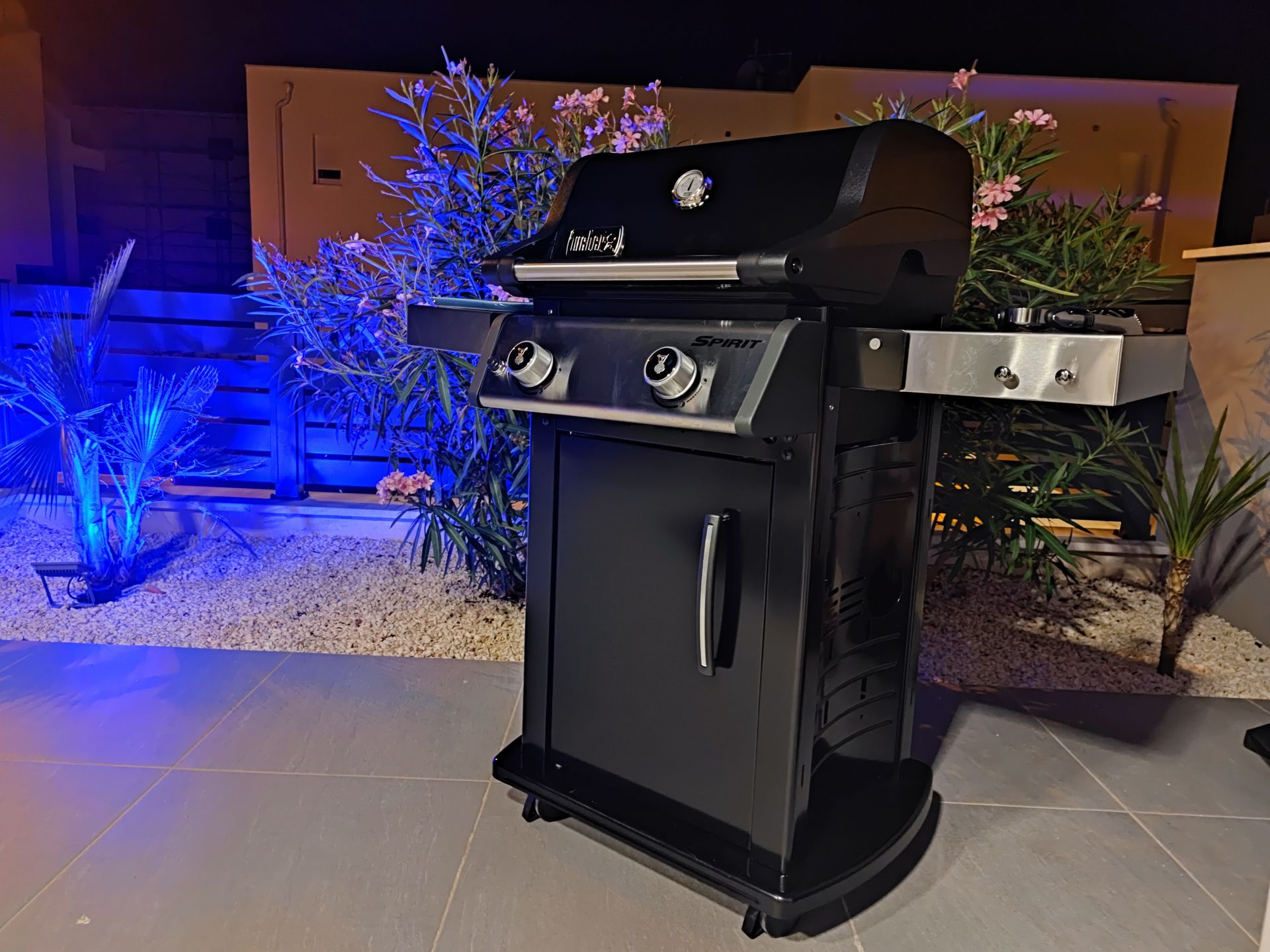 Black gas grill on a patio at night, with blue lighting and plants in the background.