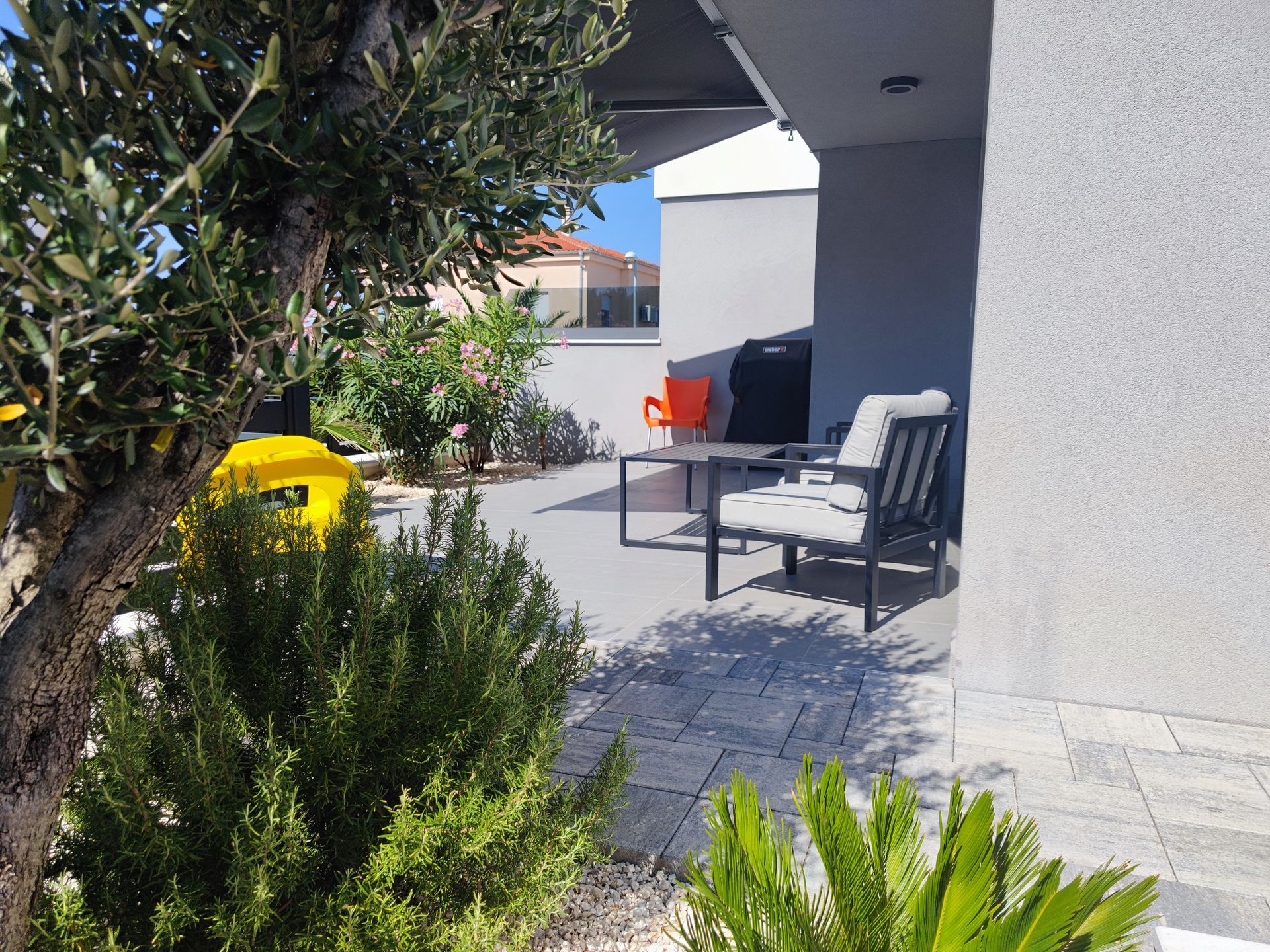 Patio with seating: a grey table and chairs, a small orange chair, with plants and a tree in the foreground.