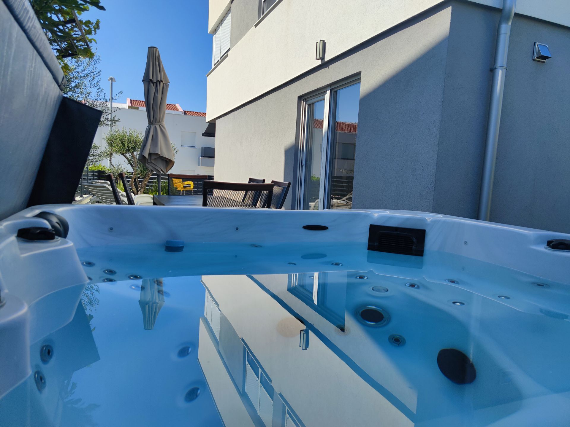 Hot tub reflecting a modern building on a sunny day. Outdoor patio with table and chairs visible.