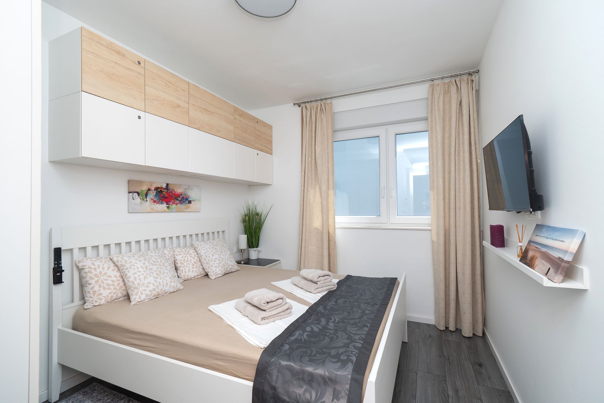 Bedroom with white bed, neutral bedding, built-in storage, TV, and window with curtains.