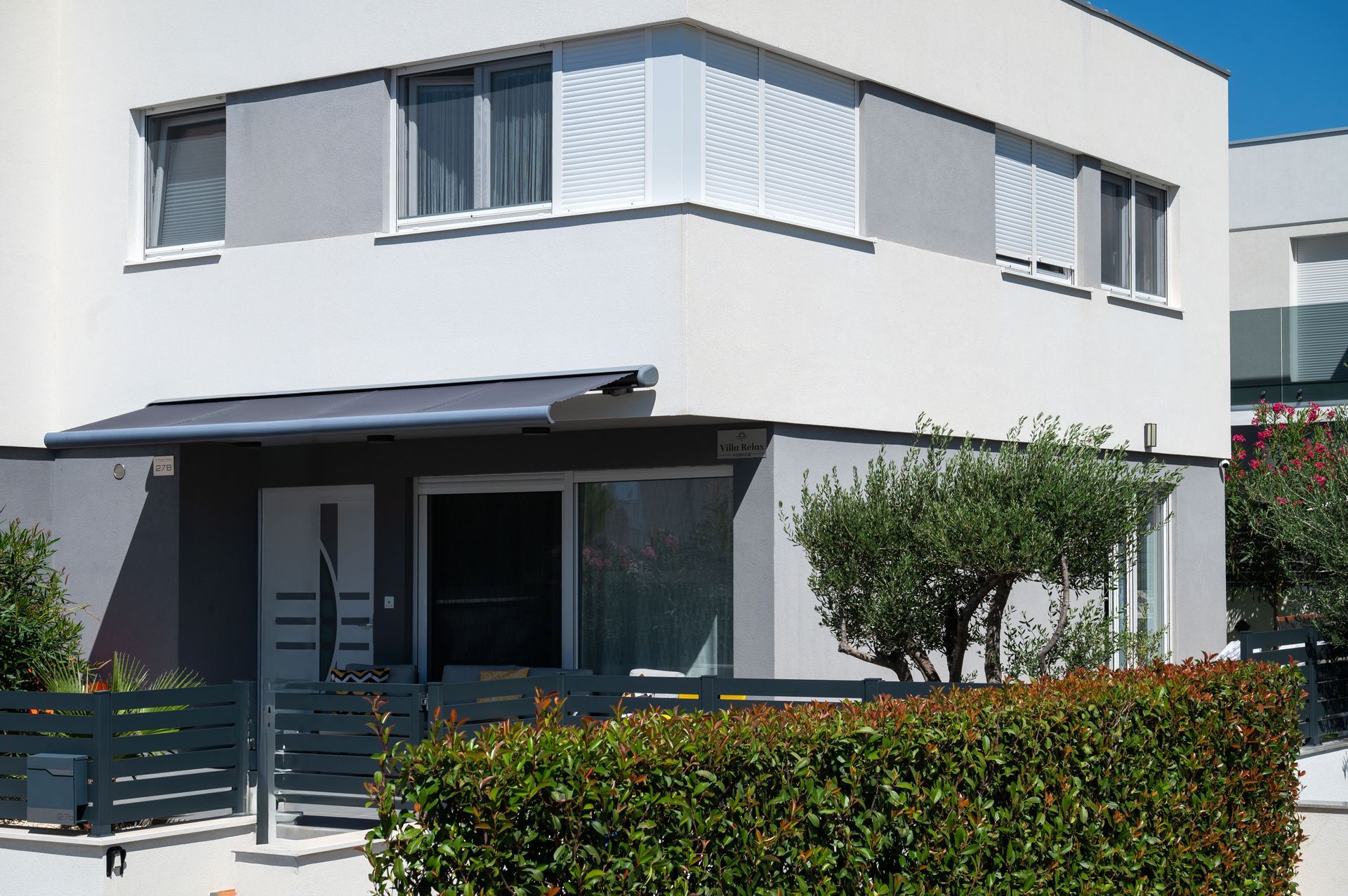 Modern two-story house with gray and white exterior, awning over the entrance, and a small hedge in front.