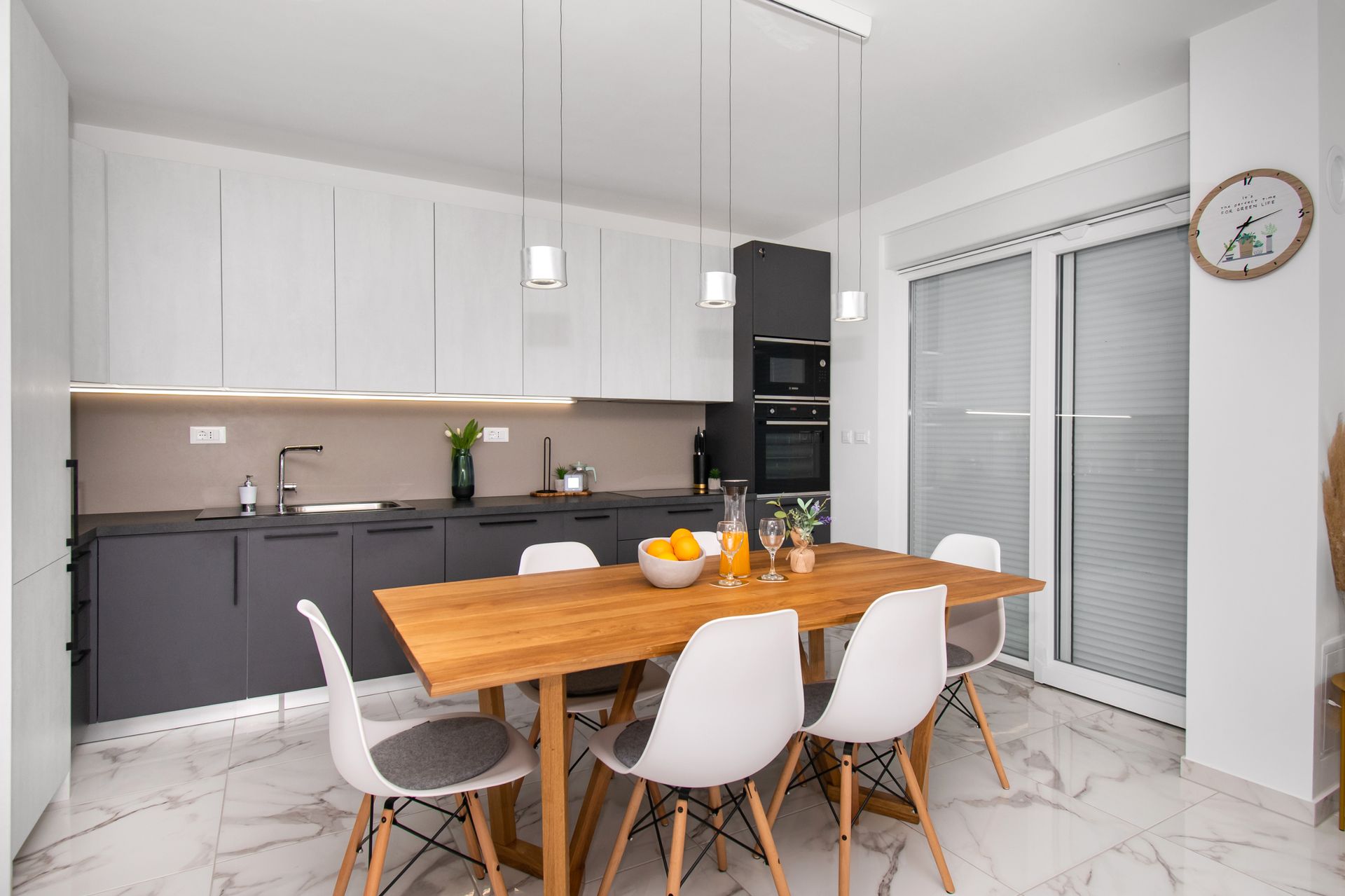 Modern kitchen with gray and white cabinets, wooden table, white chairs, and large windows.