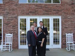 Four people pose in front of a brick building with white doors. Two rocking chairs sit nearby.