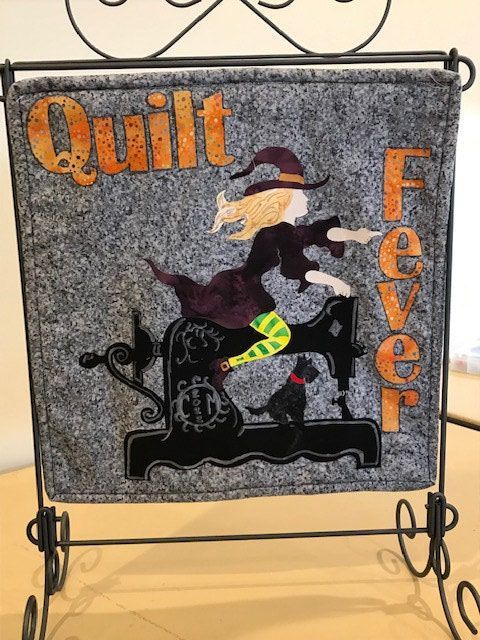 A sign that says quilt fever with a witch on a sewing machine