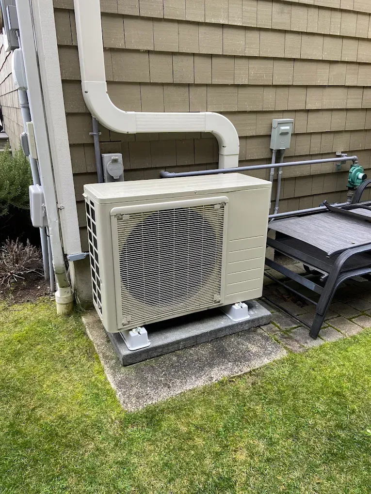 Outdoor HVAC unit on a concrete pad, against a beige house with a curved vent pipe.