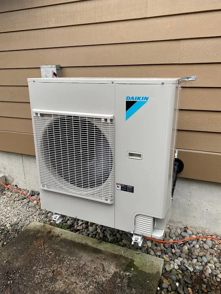 Daikin heat pump mounted on a beige building exterior, with protective grille and orange electrical wire.