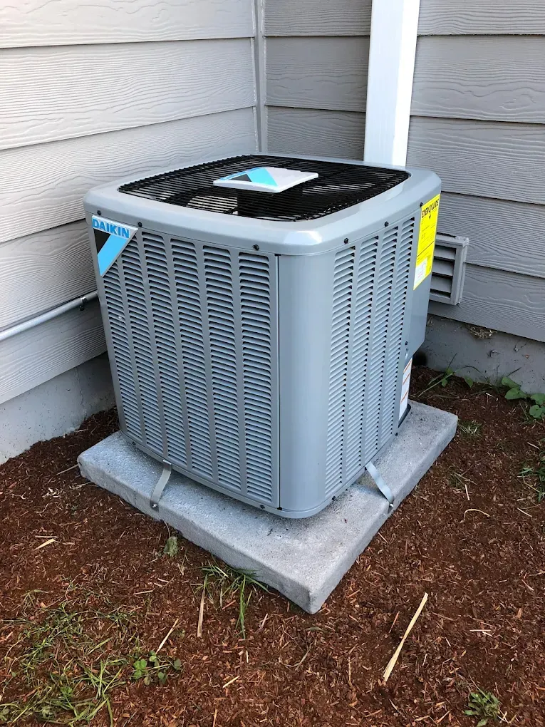 Gray air conditioner unit on concrete base beside a house with beige siding.
