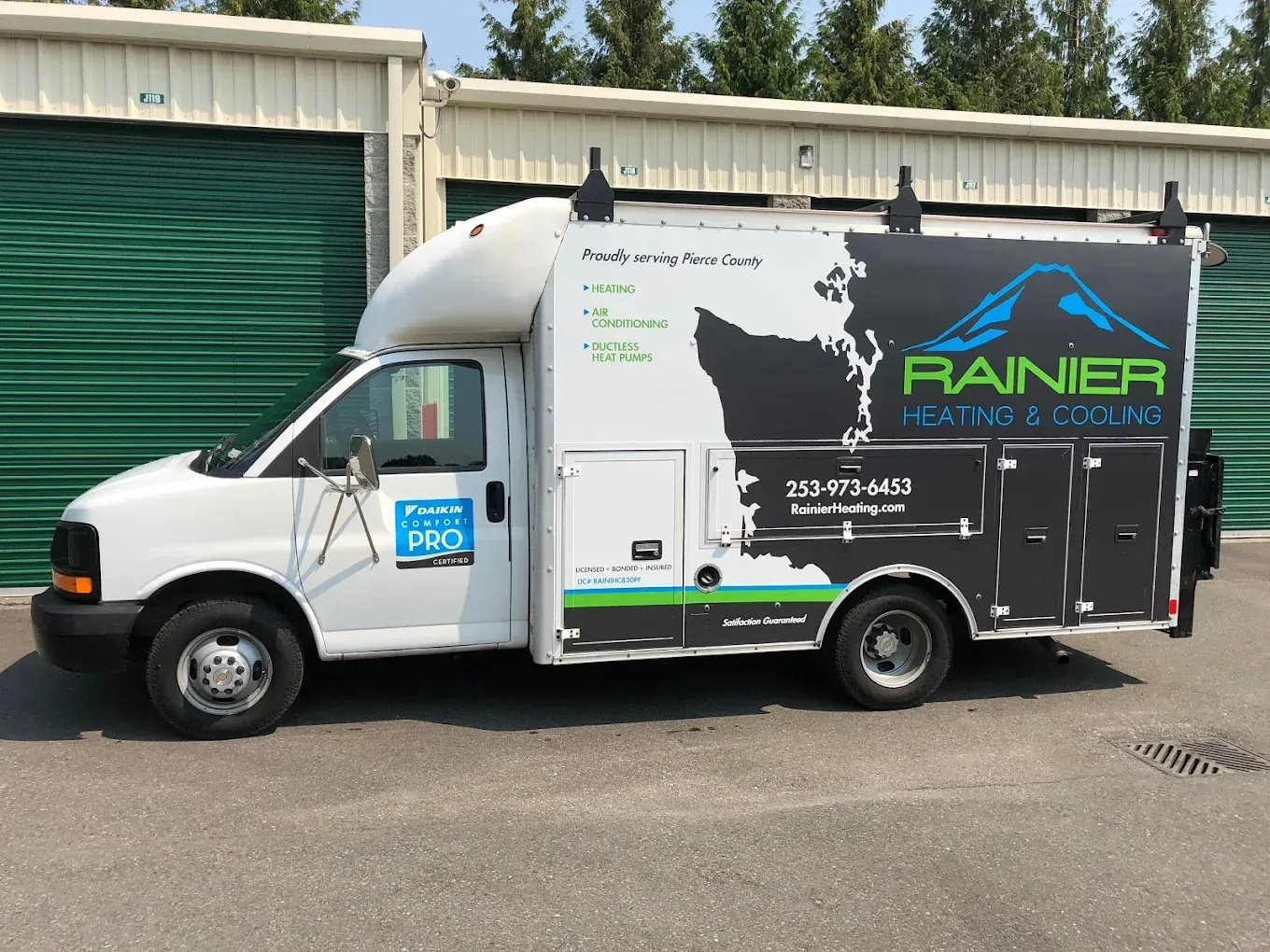 White Rainier Plumbing service van parked in front of green storage units.