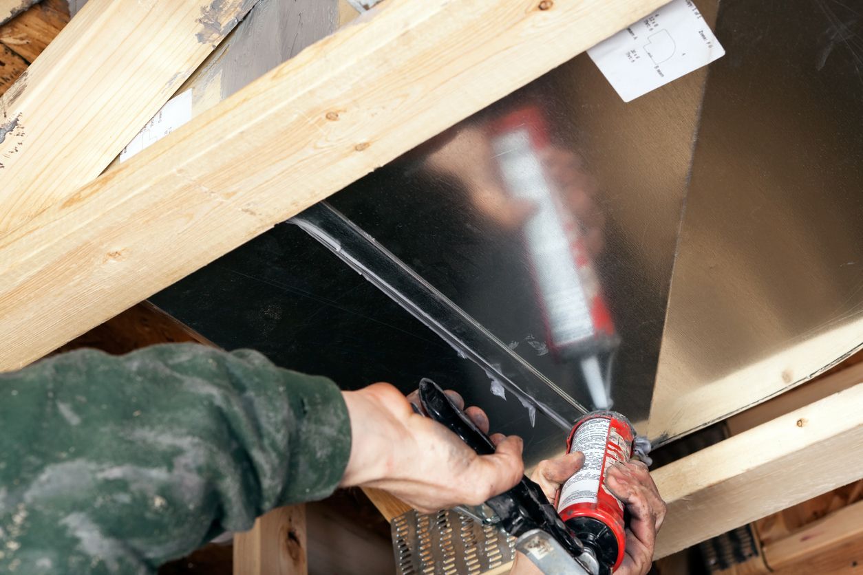 Person sealing metal sheet with caulk gun. Wooden beams support the sheet in a construction setting.