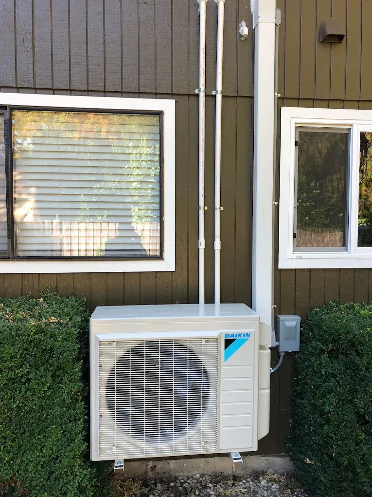 An outdoor air conditioning unit mounted on a brown wall next to windows, with white piping.
