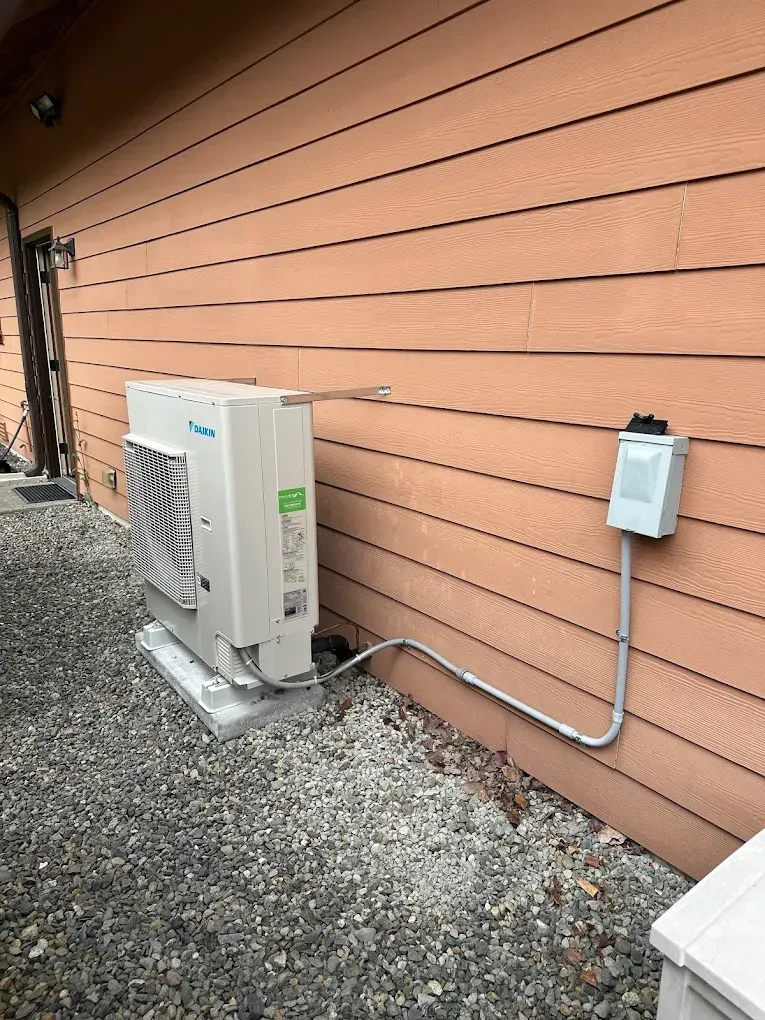 An outdoor HVAC unit on a concrete pad next to a building with brown siding. Electrical box on wall.