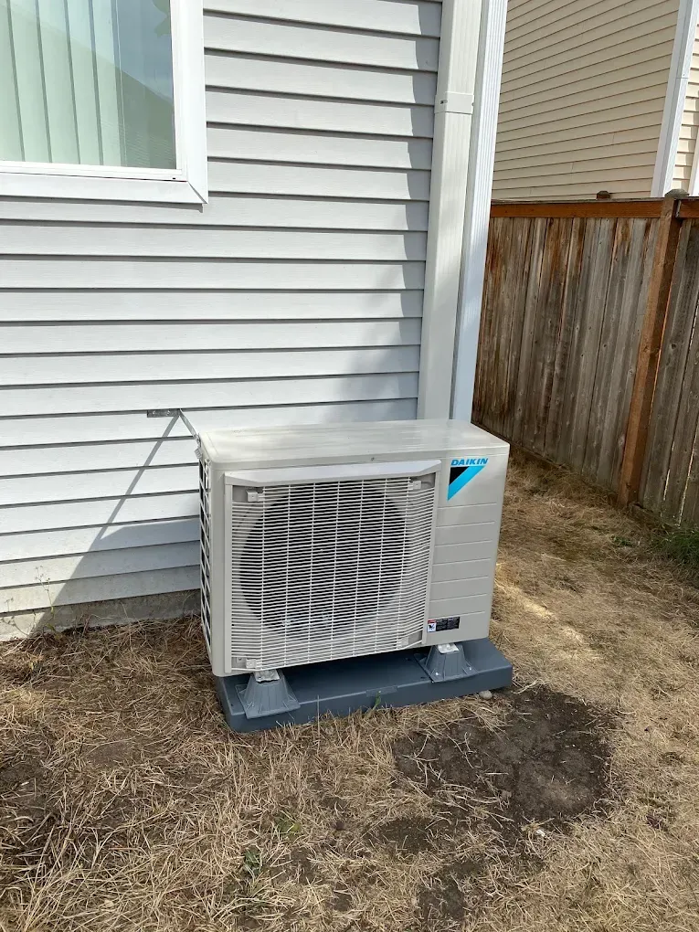 Outdoor Daikin air conditioning unit against a light gray house wall, sitting on a gray base in a grassy area.