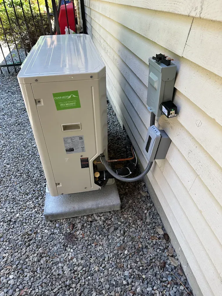 An outdoor heat pump unit next to a house wall. Gray electrical box attached with a conduit. Gravel ground.
