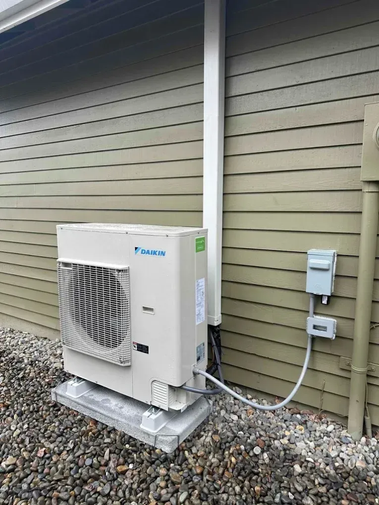 Outdoor heat pump unit next to a house with pebble ground, conduit, and electrical box.