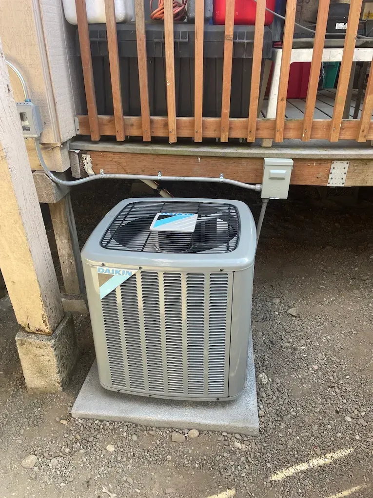 Air conditioning unit beneath a wooden deck, on a concrete pad, connected by electrical wiring.