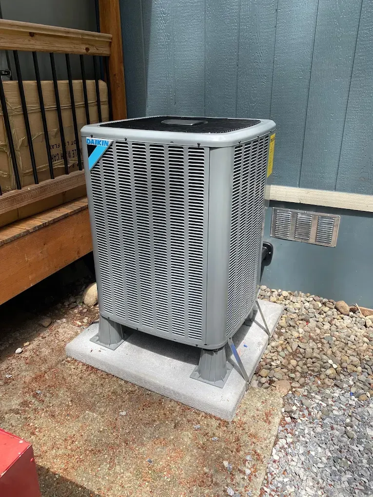 Air conditioning unit outside a building, gray and rectangular, on a concrete pad near a wooden deck.