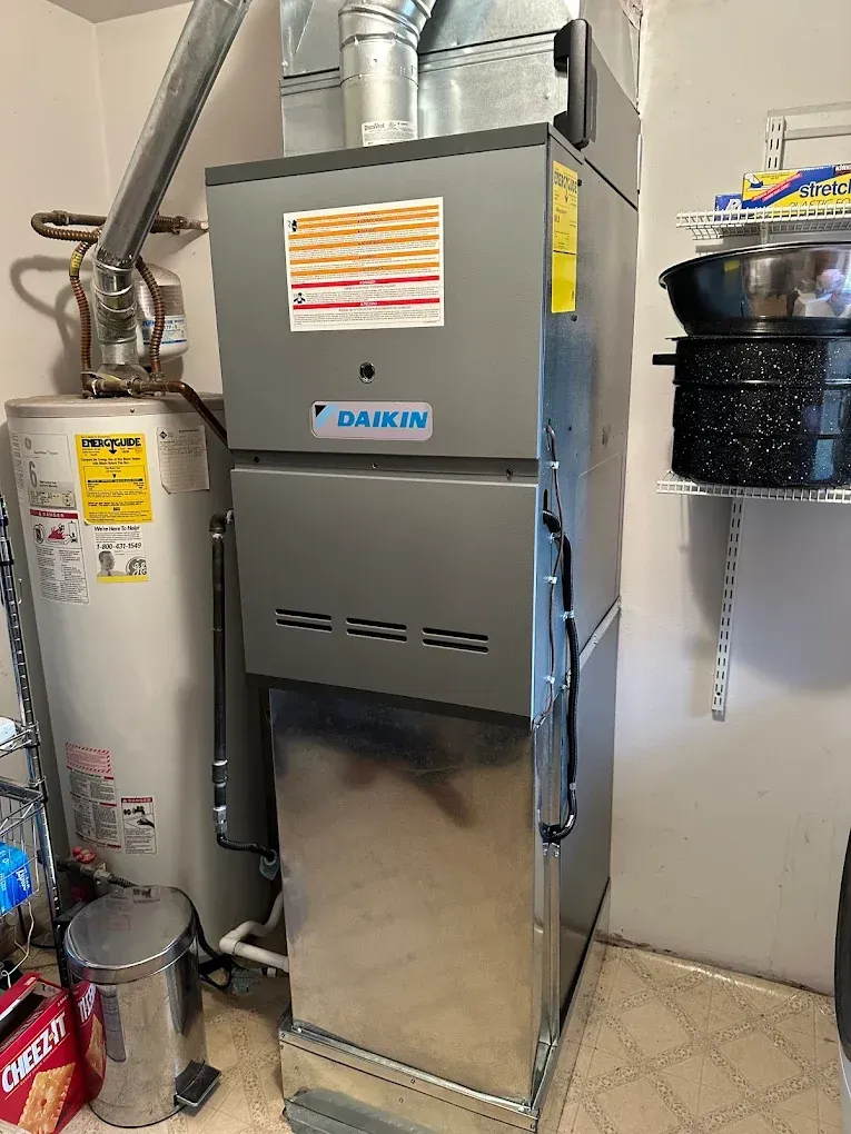Furnace and water heater in a utility room. Daikin furnace is gray with silver ductwork. Water heater is next to it.