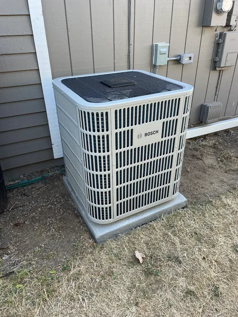 Air conditioner unit on a concrete pad next to a light-colored building with brown siding.