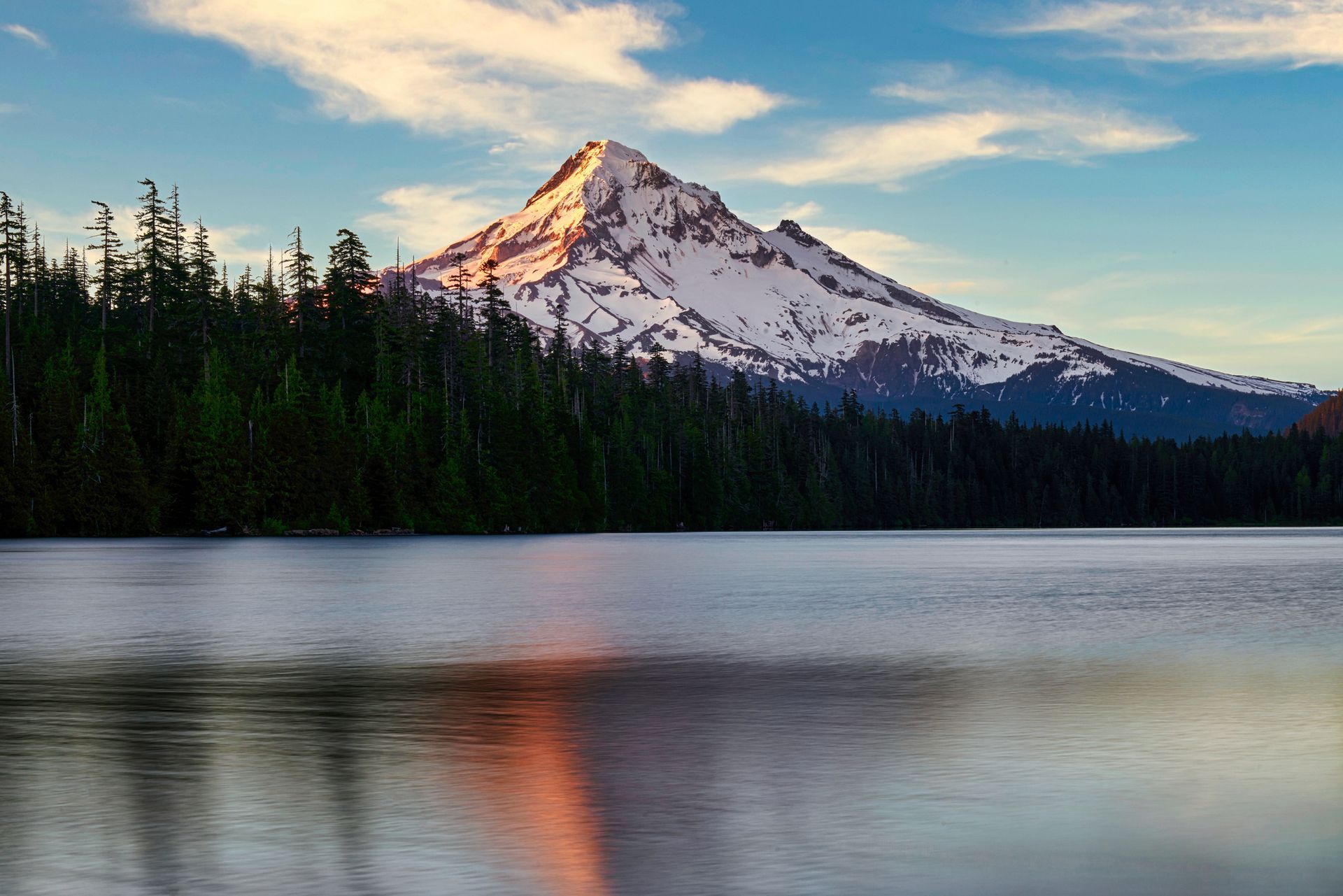 Snow-capped mountain reflecting in a calm lake, with a dark green forest along the shore under a blue sky.