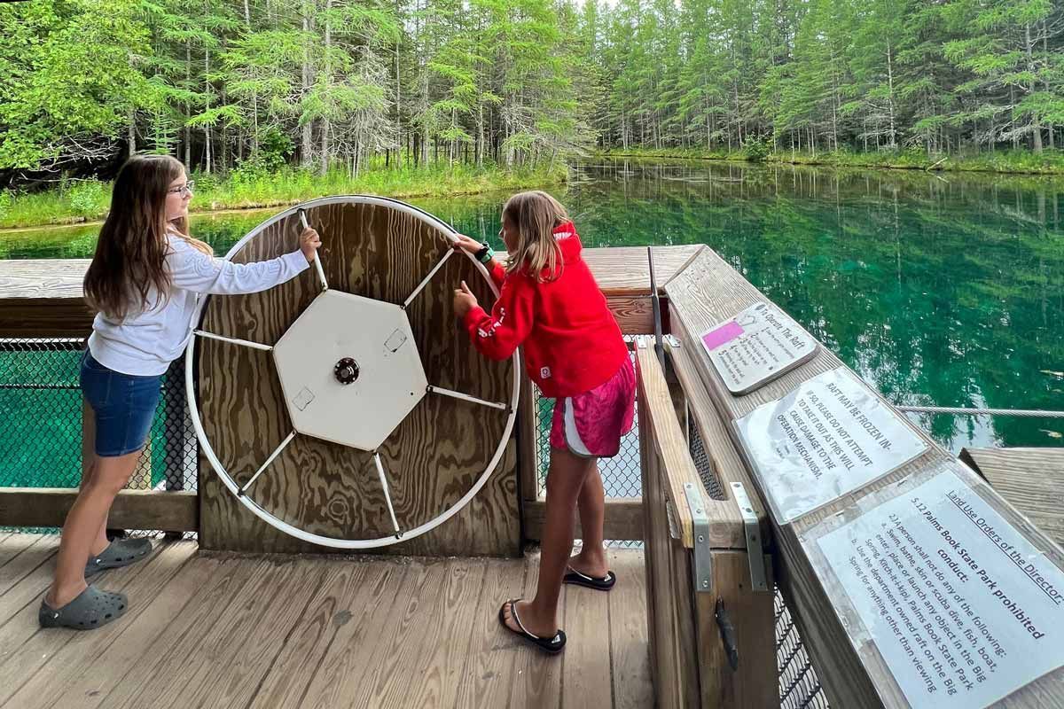 Two young girls are playing with a large wheel on a wooden deck.