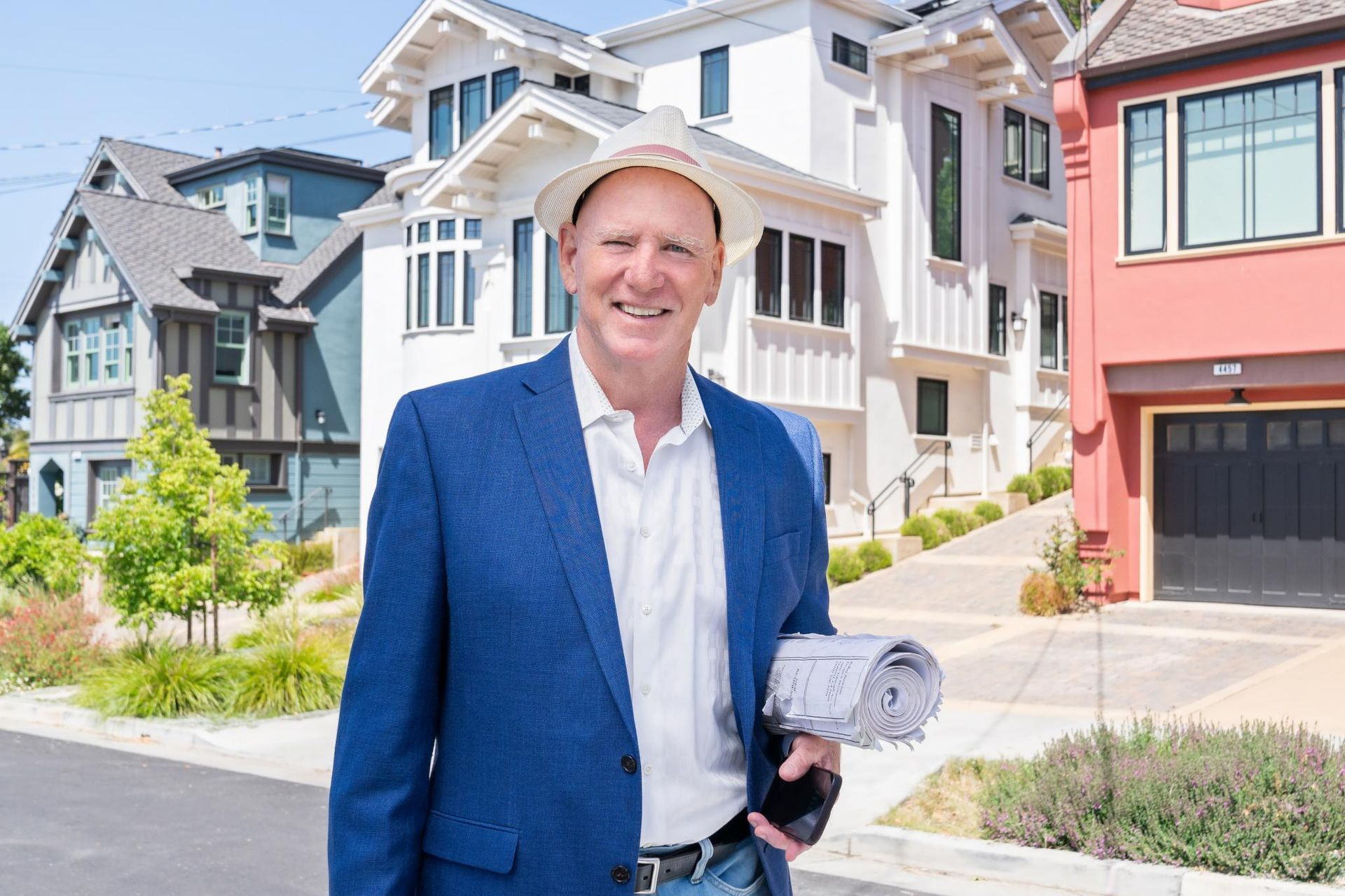 A man in a suit and hard hat is standing in front of a house holding a blueprint.