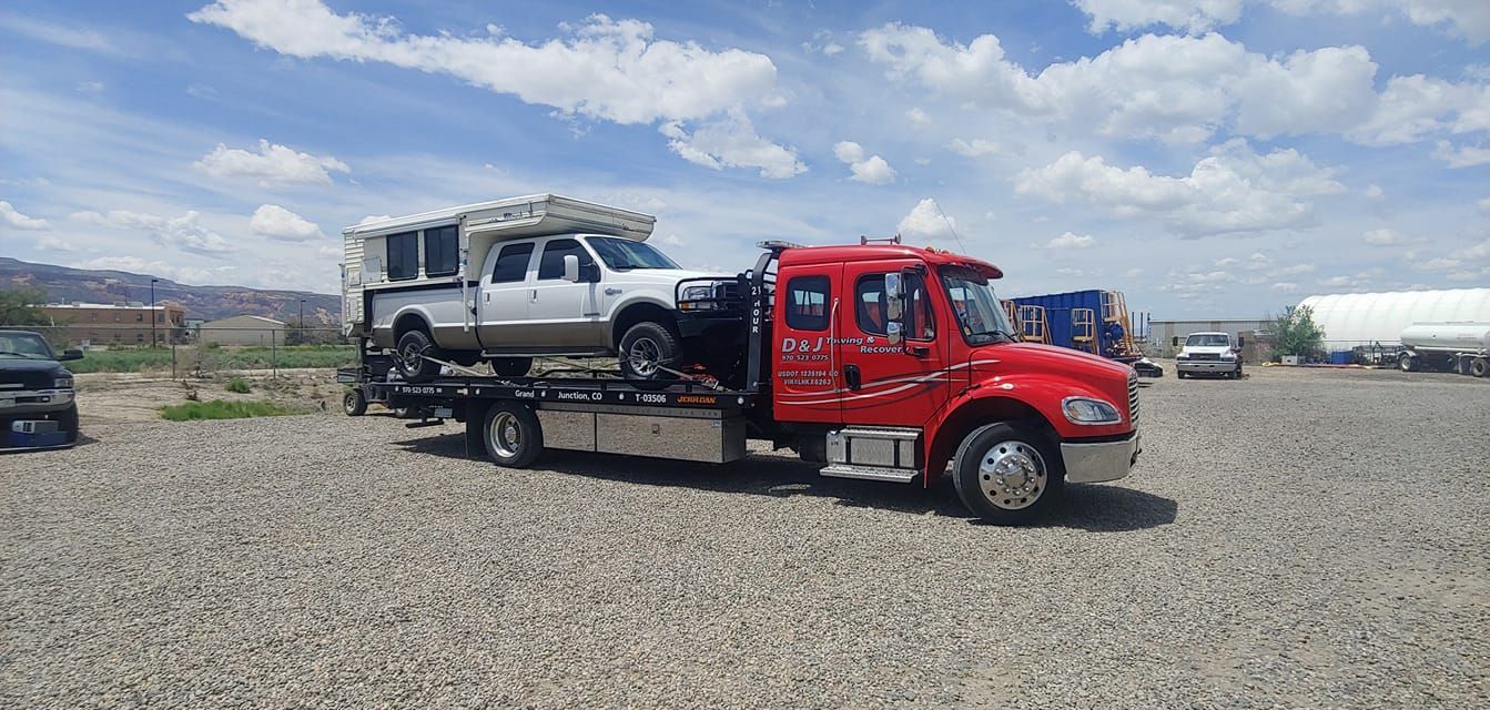 A red tow truck carrying a white pickup truck with a camper shell on a gravel lot under a cloudy sky.
