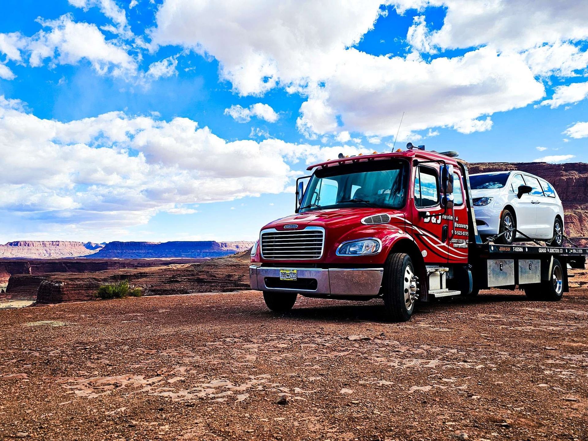 Red tow truck hauling a white car on a desert landscape, under a blue sky.