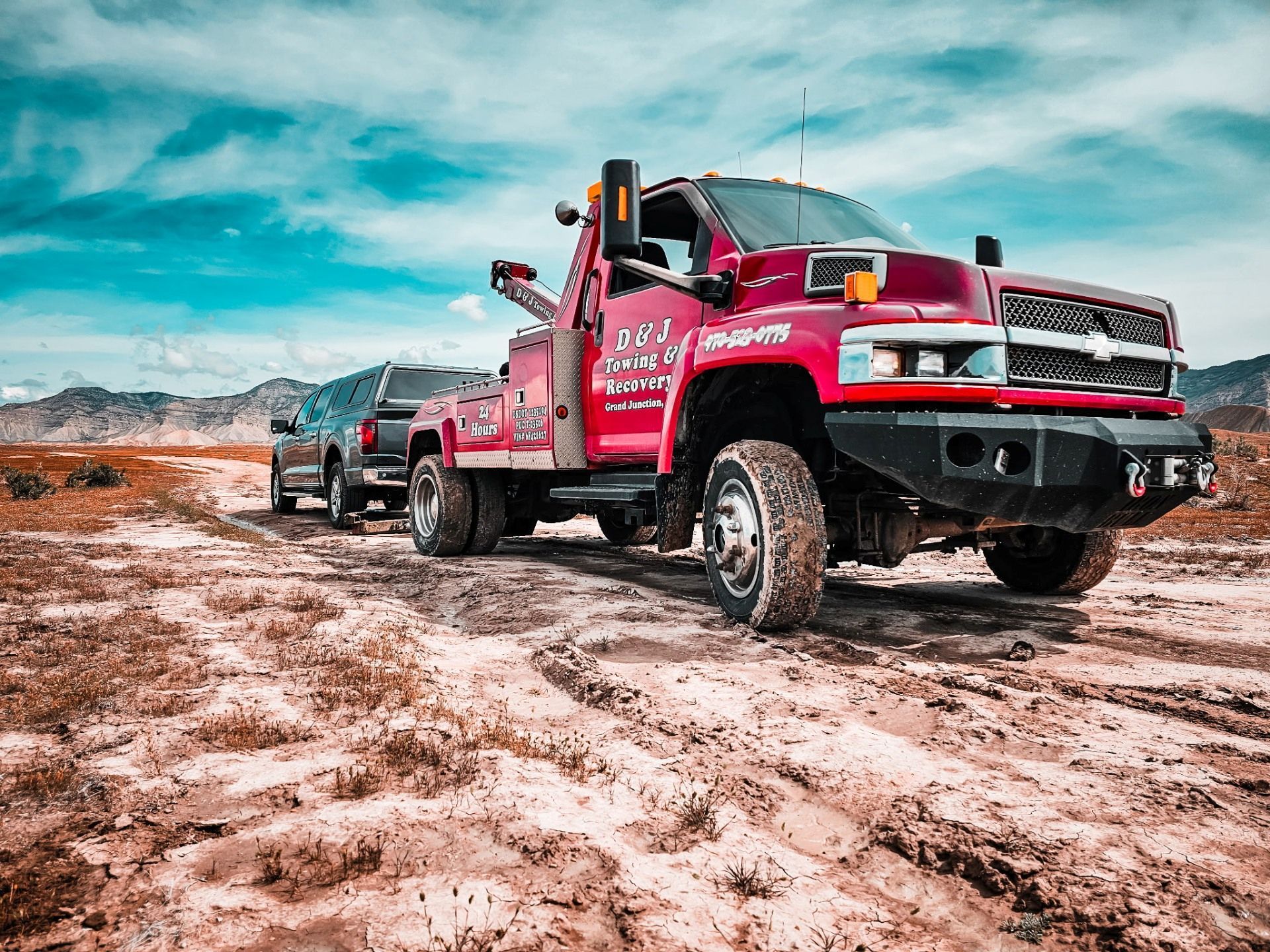 Red tow truck towing a dark truck on a dirt road under a cloudy sky.