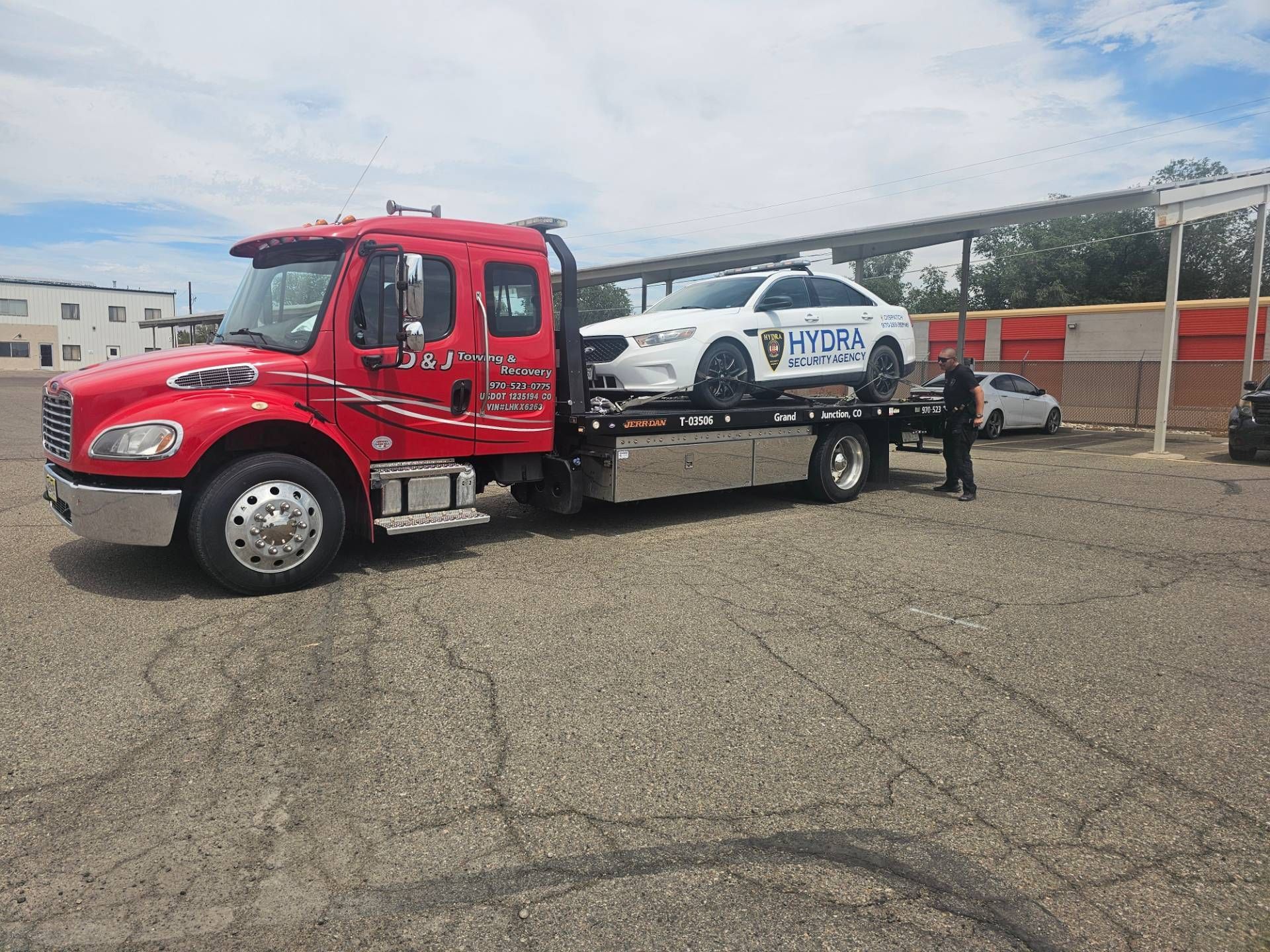 Red tow truck transporting a white police car. Setting is an asphalt lot on a sunny day.