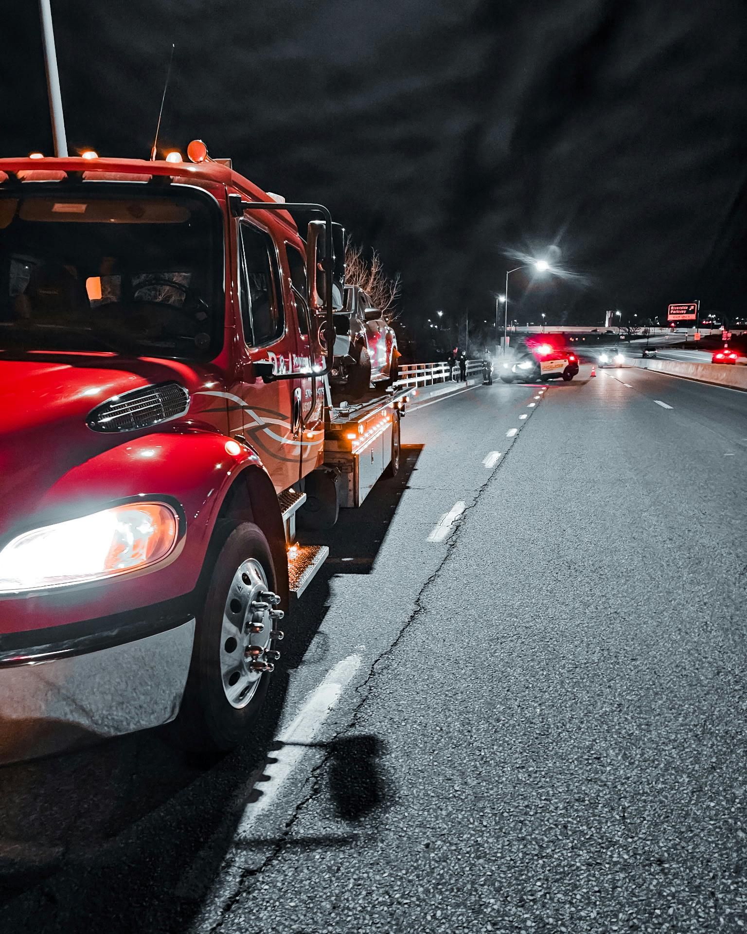 Red tow truck on a highway at night with emergency lights flashing in the distance.