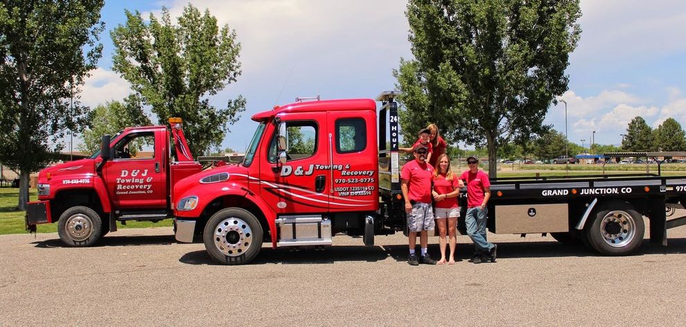 Red tow truck with three people standing beside it on a road, trees and blue sky in the background