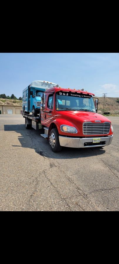 Red tow truck carrying a light blue vehicle on a paved lot on a sunny day.