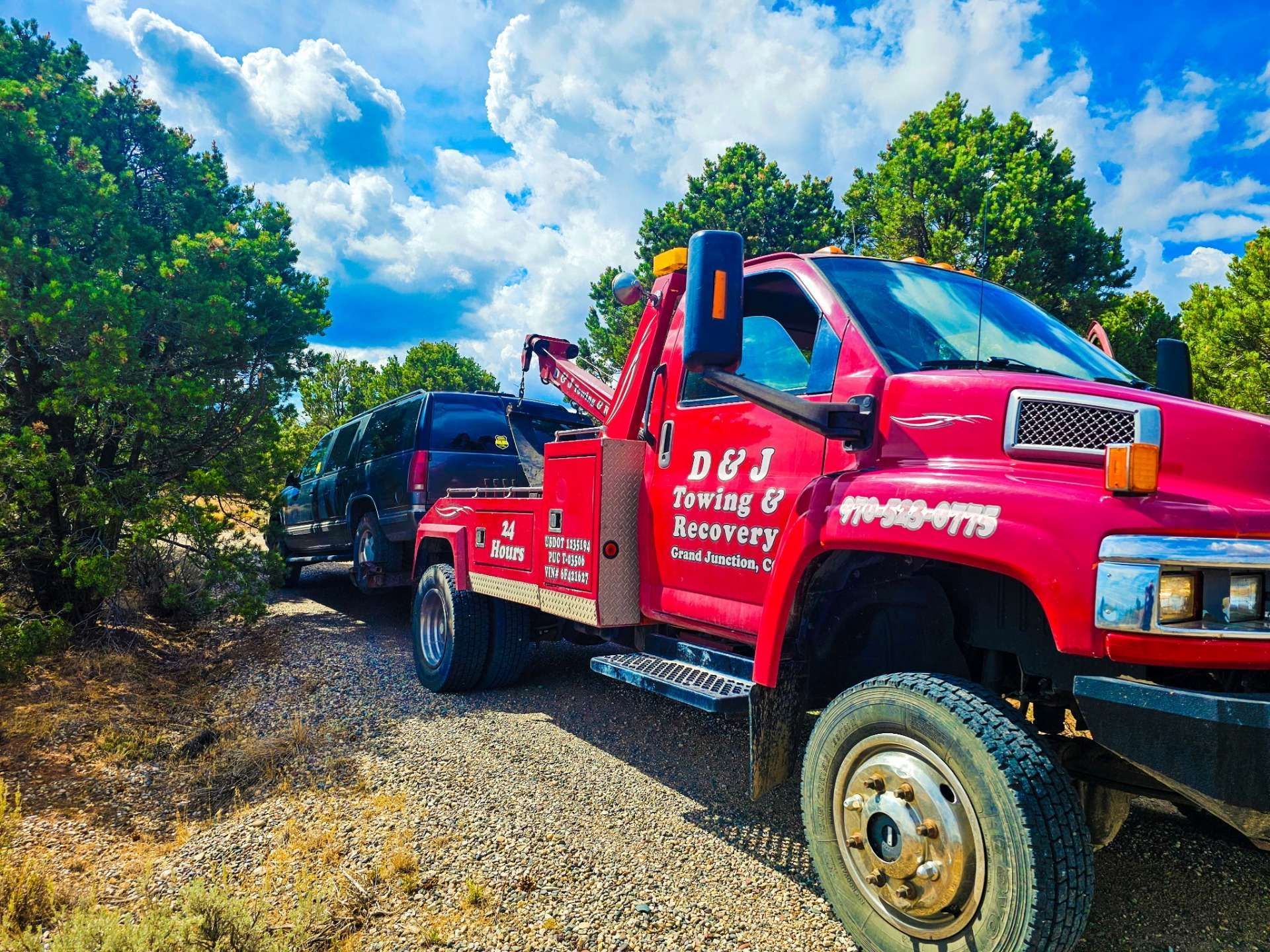 Red tow truck towing a dark vehicle on a dirt road, trees, and blue sky in the background.