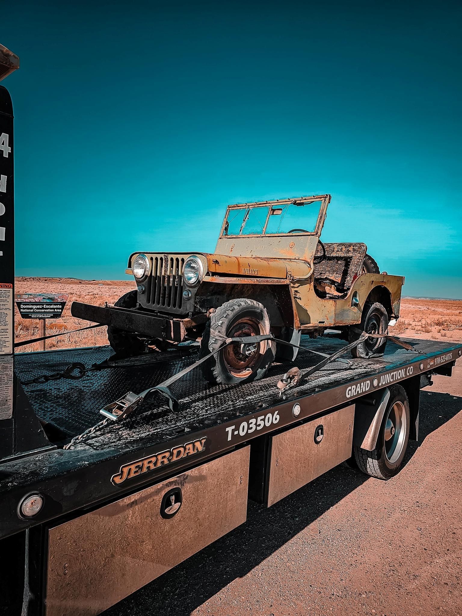 Old Jeep on a tow truck, desert landscape, clear blue sky.