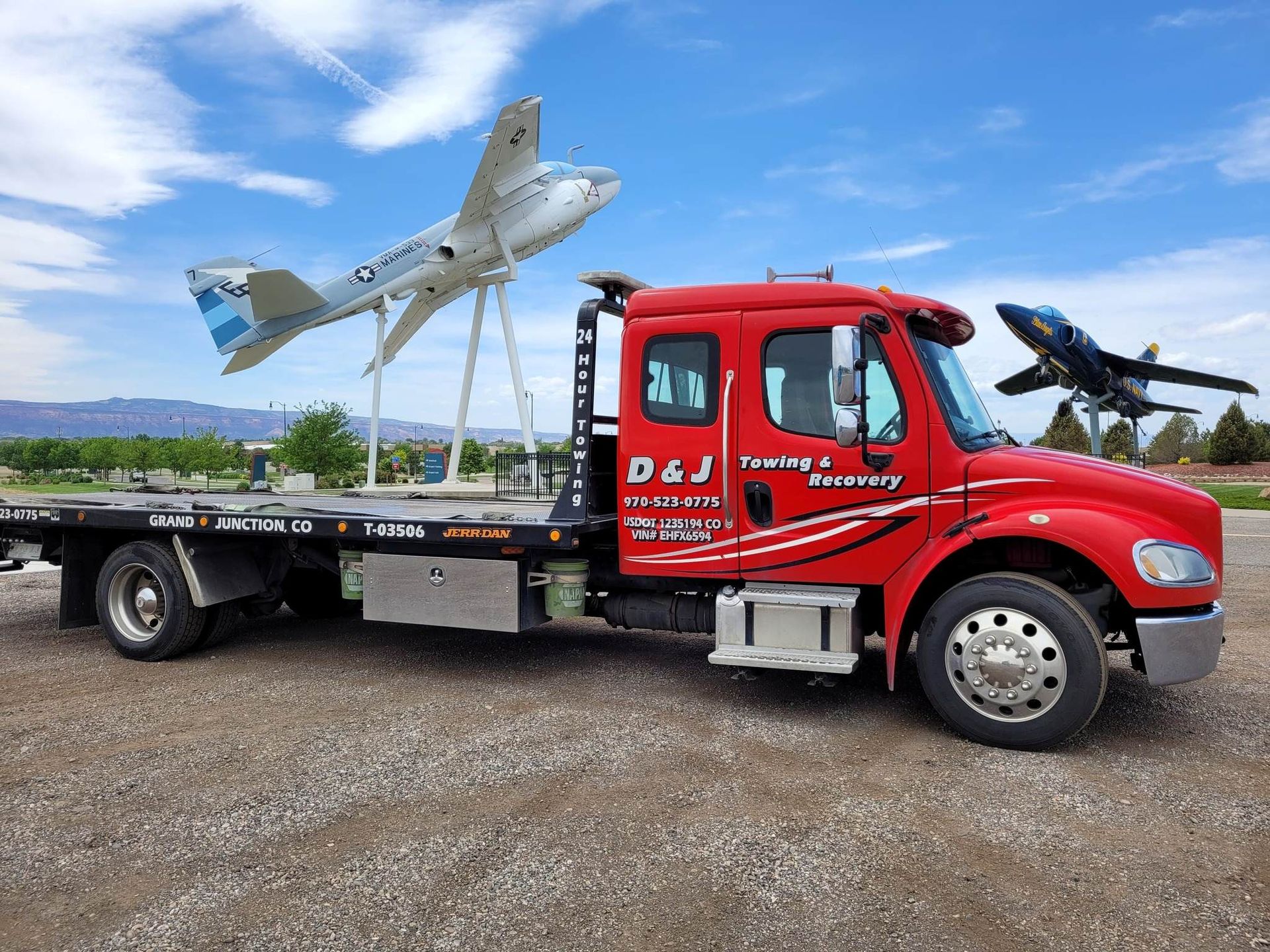 Red tow truck carrying an airplane sculpture under a blue sky, with another plane in the background.